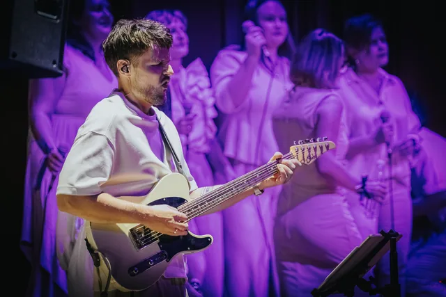 Gitarzysta grający przed chórem w fioletowym świetle.